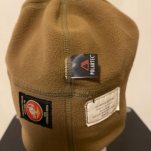 May include: A brown fleece beanie with a black "POLARTEC" label and a black patch with a gold eagle and the text "MADE EXPRESSLY FOR U.S. MARINES". The beanie also has a white label with the text "CAP HARDFACE WOVEN ELASTIC CONTRACT NO. N00140-97-C-2174 ASYLVANIA TEXTILE CO. PHILADELPHIA, PA"