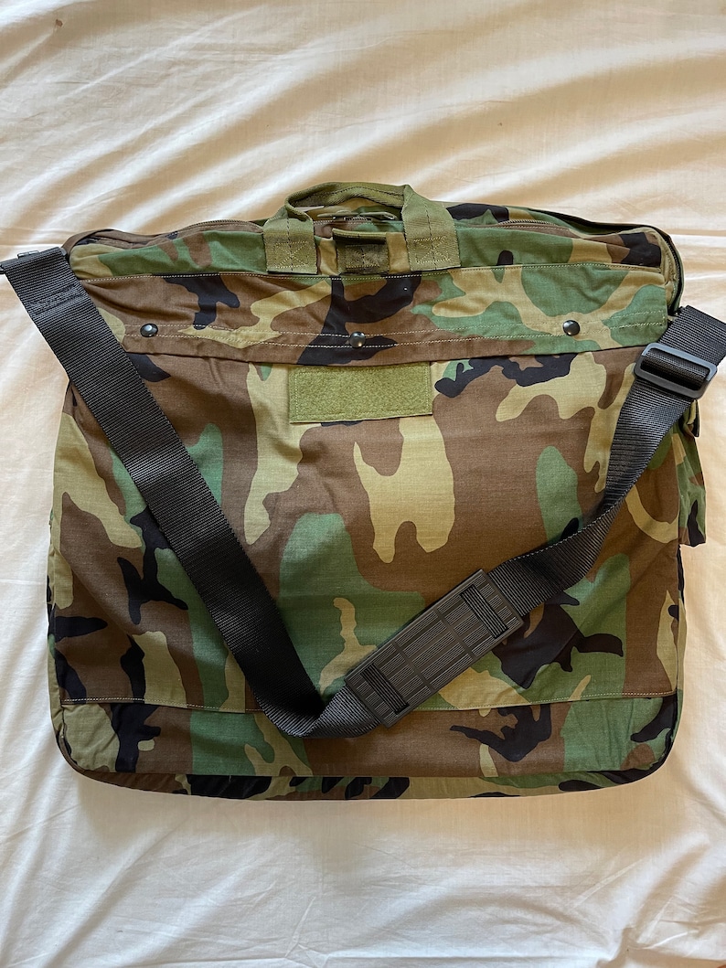 May include: A camouflage green and brown duffel bag with a black adjustable shoulder strap. The bag has a flap with a velcro closure and a small patch on the front.