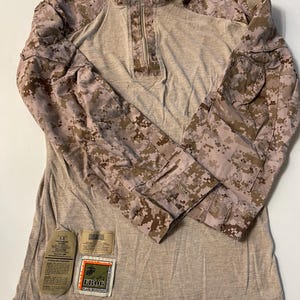 May include: A tan and brown camouflage long-sleeved shirt with a quarter-zip neckline. The sleeves and shoulders have a pixelated camouflage pattern. Patches with text are visible on the lower front. The shirt is designed for tactical use.