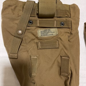 Puede incluir: Una bolsa de estilo militar de color beige con una correa grande y varias correas más pequeñas. La bolsa tiene una etiqueta que dice "MAGAZINE, M16, 30 ROUND, NSN 1005-01-515-5703, LOT NO. 99CT-57M22, NSN 8465-01-584-0054, MFG. WHITMAN FABRICATING."