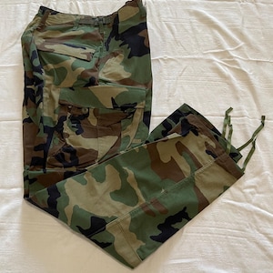 Könnte beinhalten: Tarnfarbene Cargohose im Waldmuster mit Grün-, Braun- und Schwarztönen. Die Hose hat mehrere Taschen, einen Knopfverschluss und Kordelzüge an den Knöcheln. Der Stoff scheint ein strapazierfähiges Material zu sein.