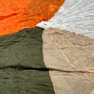 May include: A colorful parachute with segments of orange, olive green, tan, and white fabric. The parachute is spread out, showing the texture and pattern of the material. The image is taken from above, highlighting the circular design.