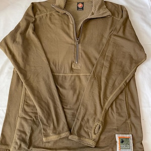 May include: A tan long-sleeved shirt with a quarter-zip closure and a small collar. The shirt has a textured pattern and a small patch on the lower right side with the text "FROG". The shirt has thumb holes at the cuffs.