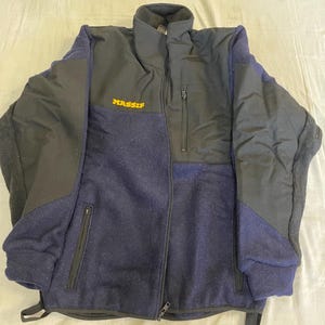 May include: A navy blue and black zip-up jacket featuring a high collar, long sleeves with ribbed cuffs, and two lower zippered pockets. The word MASSIF is printed in yellow on the upper right chest, and a vertical zippered pocket is on the left chest.