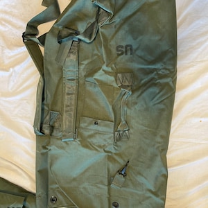 May include: A green canvas duffel bag with a black strap and a black "US" logo. The bag has a zipped pocket and a handle.