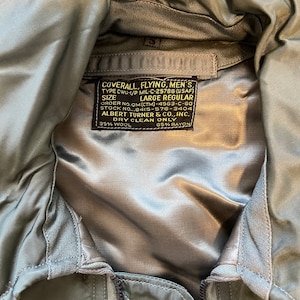May include: A close-up of a sage green flying jacket. The jacket has a satin-like lining and a label with text including "COVERALL, FLYING, MEN'S" and "LARGE REGULAR". The jacket has a zipper and quilted stitching.