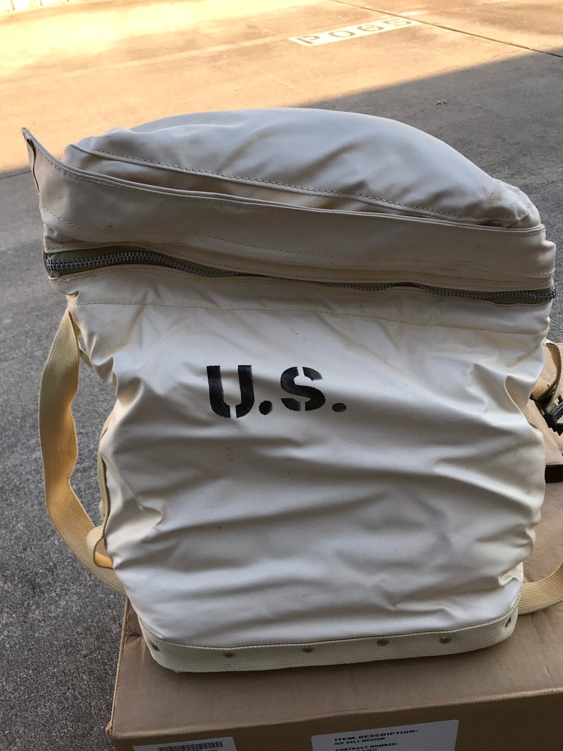 May include: A white canvas bag with a zipper closure and a black "U.S." stencil. The bag has a brown leather strap and a white canvas strap.