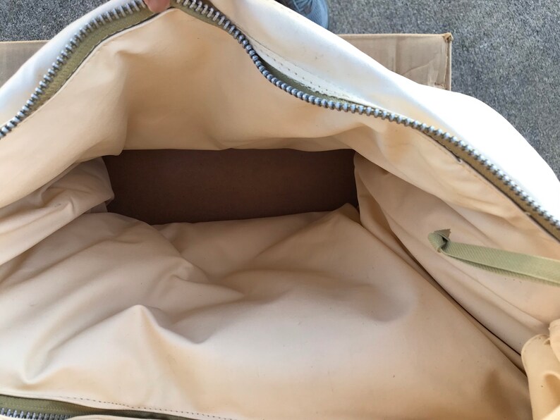 May include: A white canvas duffel bag with a green zipper and a green strap. The bag is open and empty.