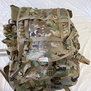 Pode incluir: Mochila com padrão de camuflagem com várias correias, fivelas e compartimentos. A mochila apresenta uma mistura de cores verdes, castanhas e bege. Possui correias ajustáveis e um fecho de aba. Uma pequena etiqueta com um código de barras é visível.