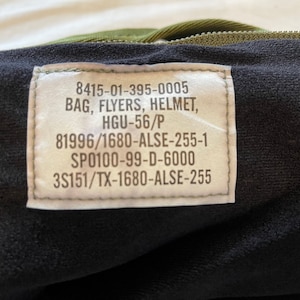 May include: A close-up of a green military bag with a label that reads "8415-01-395-0005 BAG, FLYERS, HELMET, HGU-56/P 81996/1680-ALSE-255-1 SP0100-99-D-6000 3S151/TX-1680-ALSE-255".