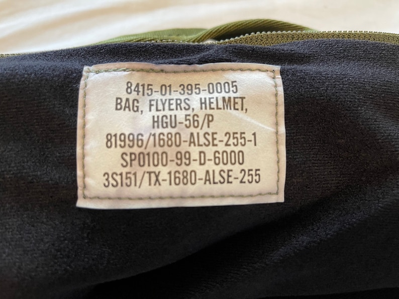May include: A close-up of a green military bag with a label that reads "8415-01-395-0005 BAG, FLYERS, HELMET, HGU-56/P 81996/1680-ALSE-255-1 SP0100-99-D-6000 3S151/TX-1680-ALSE-255".