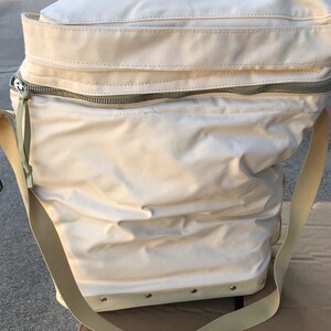 May include: A white canvas bag with a zipper closure and a long strap. The bag has a reinforced bottom with metal rivets.