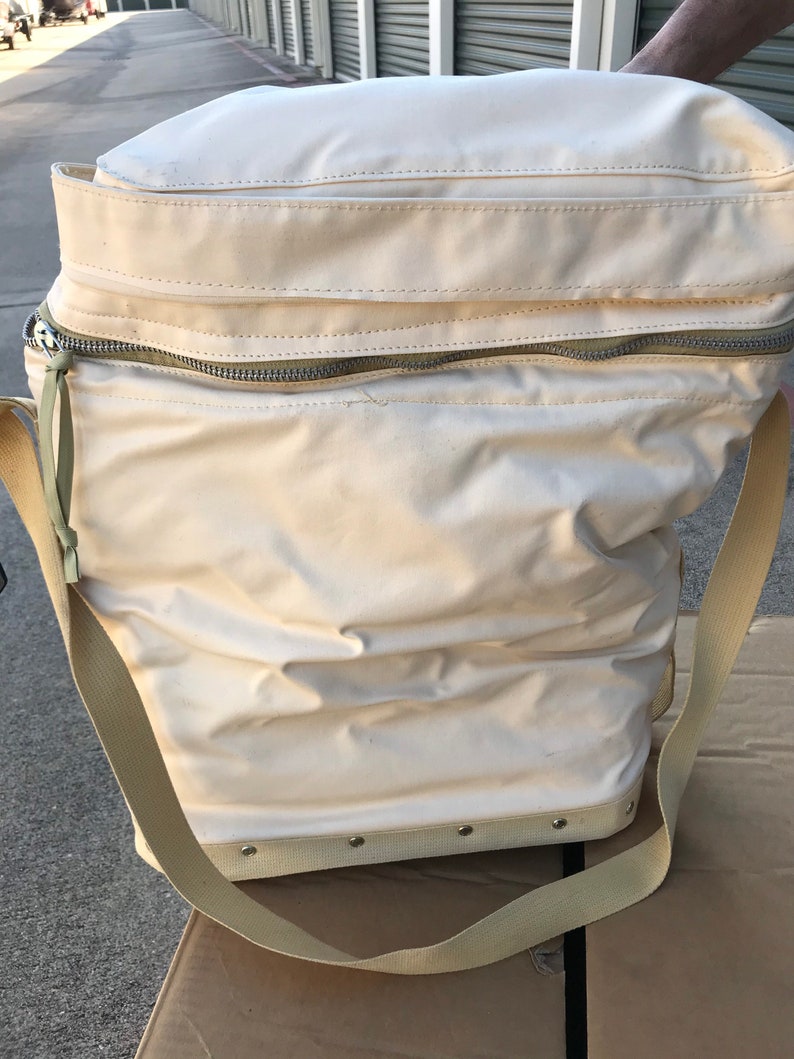 May include: A white canvas bag with a zipper closure and a long strap. The bag has a reinforced bottom with metal rivets.
