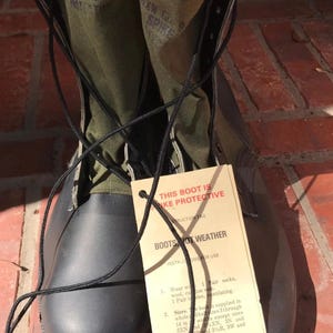 May include: A pair of black rubber boots with olive green canvas uppers and black laces. A tan instruction tag is attached, reading: THIS BOOT IS SPIKE PROTECTIVE, BOOTS NOT WEATHER, INSTRUCTIONS FOR USE, 1. Wear with 1 pair socks, wool, cushion sole, 1 pair insole, ventilating.