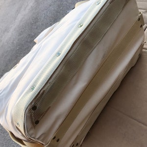May include: A white canvas tote bag with reinforced seams and multiple straps. The bag has a cream colored canvas trim and is secured with metal rivets.