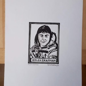 以下が含まれることがあります： イギリス初の宇宙飛行士、ヘレン・シャーマンの白黒リノカットプリント。宇宙服を着たシャーマンの肖像画が特徴で、下に「HELEN SHARMAN」という名前が印刷されています。アーティストのサイン入り。