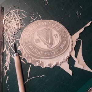 Op de afbeelding: Een uitgesneden rubberen stempel van een Guinness-flesdopontwerp, met het harp-logo en tekst. De stempel ligt op een groene snijmat, met een snijgereedschap en verwijderde rubberen stukken in de buurt.