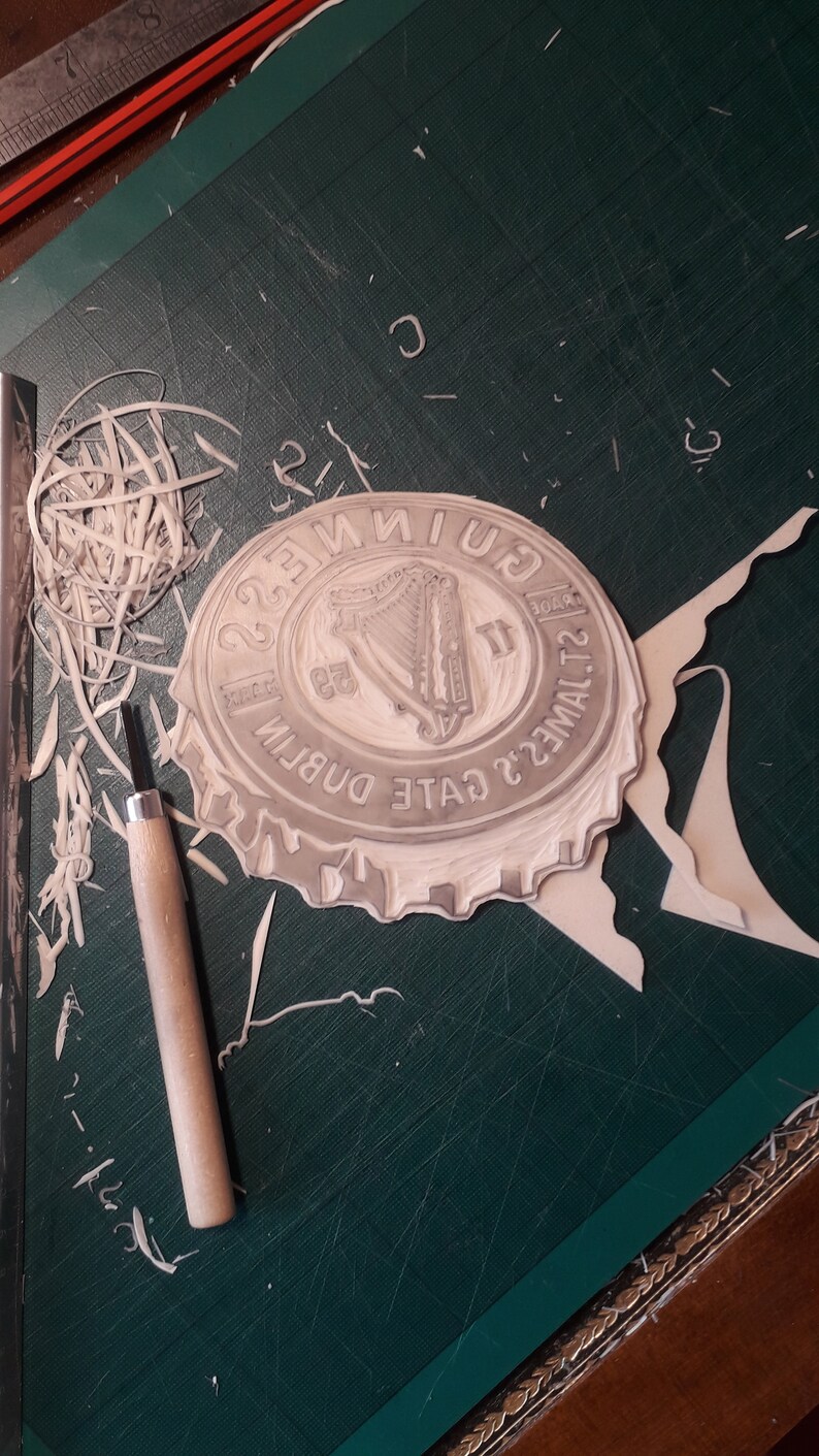 Op de afbeelding: Een uitgesneden rubberen stempel van een Guinness-flesdopontwerp, met het harp-logo en tekst. De stempel ligt op een groene snijmat, met een snijgereedschap en verwijderde rubberen stukken in de buurt.