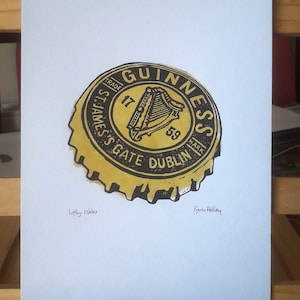 Op de afbeelding: Een print van een Guinness-flesdop. De dop is geel en zwart met het Guinness-logo en de tekst. De print staat op wit papier. De tekst "Liffey Water" en de handtekening van de kunstenaar zijn ook zichtbaar.