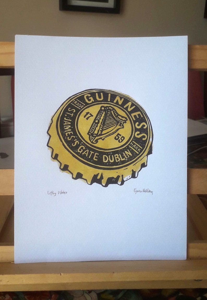 Op de afbeelding: Een print van een Guinness-flesdop. De dop is geel en zwart met het Guinness-logo en de tekst. De print staat op wit papier. De tekst "Liffey Water" en de handtekening van de kunstenaar zijn ook zichtbaar.