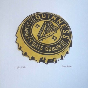 Op de afbeelding: Een afdruk van een Guinness-flesdop. De dop is geel en zwart, met het Guinness-logo en tekst. De tekst "St. James's Gate Dublin" is zichtbaar. De afdruk bevat ook de tekst "Liffey Water" en de handtekening van de kunstenaar.