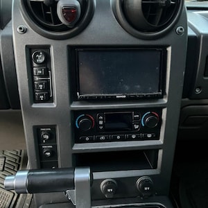 May include: A gray car dashboard with a black Kenwood radio, a black and gray gear shifter, and two 12V power outlets. The radio has a screen, buttons, and knobs. The gear shifter has a black handle. The power outlets are labeled "12V".