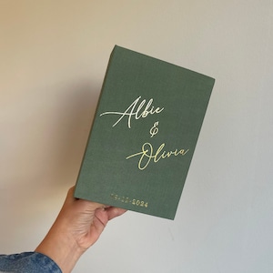 May include: A dark green guest book with the names "Albie & Olivia" written in white script and the date "05.12.2024" in gold lettering.