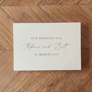 May include: A white card with gold lettering that reads "Our Wedding Day" followed by the names "Rebecca and Brett" and the date "23 March 2018".