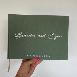 以下が含まれることがあります： 白の筆記体で「Brandon and Elyse」と書かれた緑色のハードカバー本。日付「18th January 2024」が名前の下に印刷されています。薄緑色のリボンブックマークが付いています。