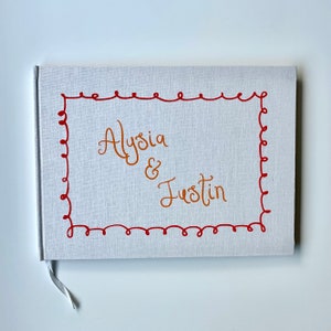 Puede incluir: Un libro blanco cubierto de lino con los nombres "Alysia & Justin" en escritura naranja. Los nombres están dentro de un borde rojo con líneas onduladas. Una cinta blanca sirve como marcapáginas.