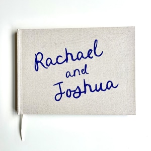 Op de afbeelding: Een boek met linnen omslag met de namen "Rachael en Joshua" in blauw schrift. Het boek heeft een crèmekleurige kaft en een wit lint als bladwijzer. De namen zijn geschreven in een stijlvol, cursief lettertype.