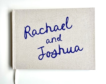 Puede incluir: Un libro con cubierta de lino con los nombres "Rachael y Joshua" en escritura azul. El libro tiene una cubierta de color crema y una cinta blanca como marcador. Los nombres están escritos en una fuente cursiva elegante.