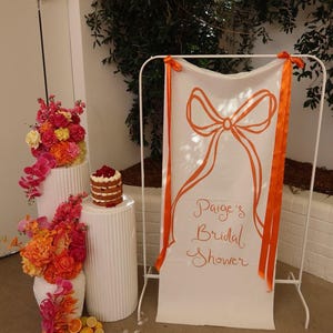 May include: A white banner with orange ribbon ties and the text "Paige's Bridal Shower" in orange cursive writing. The banner is hanging on a white stand. There are two white pillars with orange and pink flowers on them. A three-tiered cake is on the top of one of the pillars.