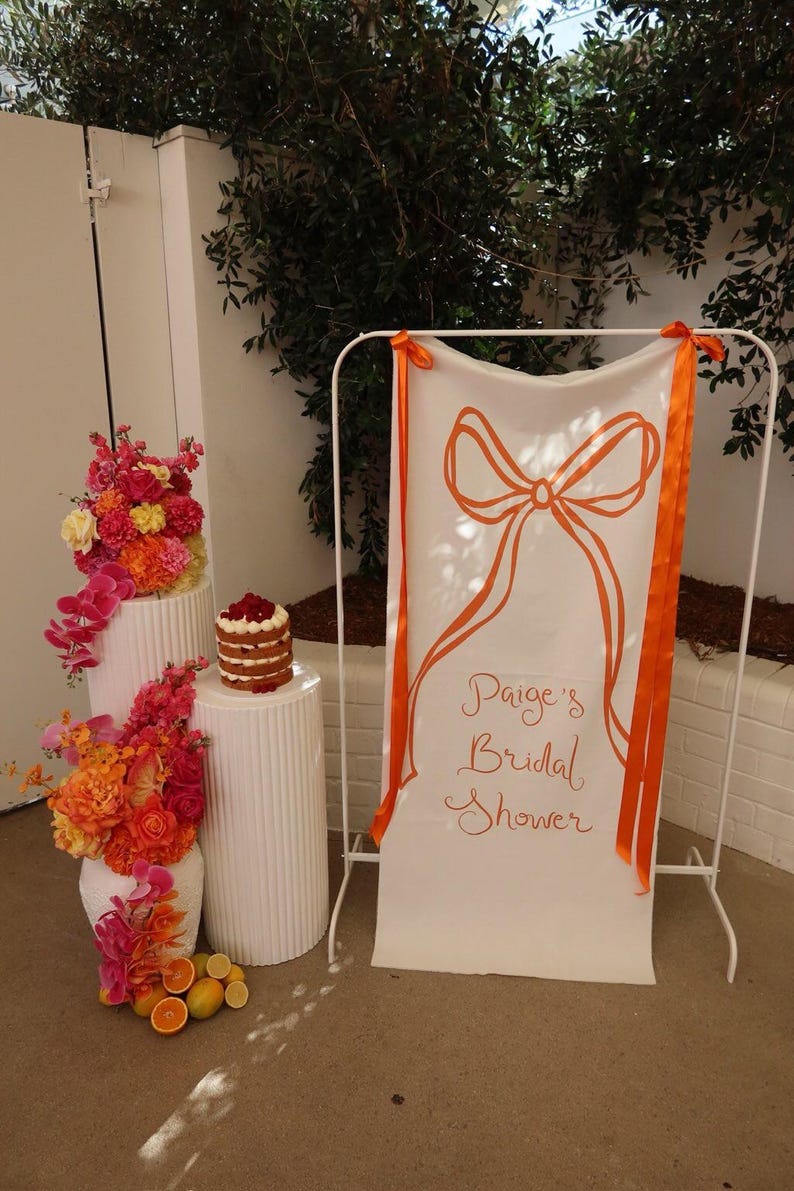 May include: A white banner with orange ribbon ties and the text "Paige's Bridal Shower" in orange cursive writing. The banner is hanging on a white stand. There are two white pillars with orange and pink flowers on them. A three-tiered cake is on the top of one of the pillars.