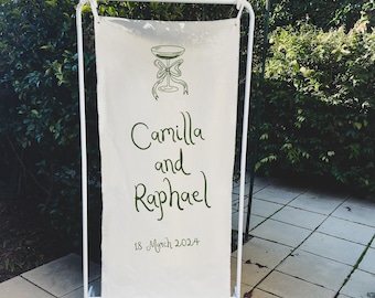 May include: White sign with a green illustration of a champagne flute with a bow on a white stand. The sign reads "Camilla and Raphael 18 March 2024".