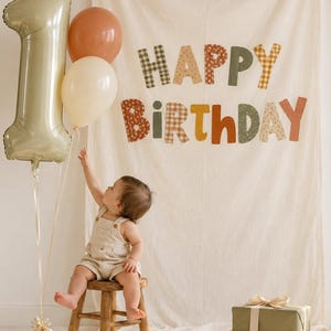 May include: A cream-colored fabric backdrop featuring the text HAPPY BIRTHDAY in multi-colored patterned letters, accompanied by a large number one balloon, two round balloons, a wooden stool, and two stacked olive green gift boxes with ribbons.