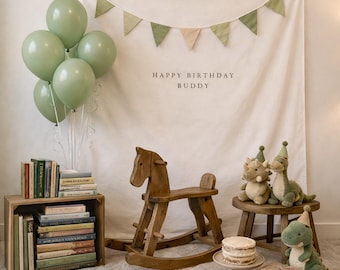 May include: A birthday party setup featuring a cream-colored backdrop with green bunting and the text HAPPY BIRTHDAY BUDDY. Includes sage green balloons, a wooden rocking horse, a stack of books, three plush dinosaurs, and a cake.