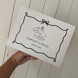 May include: A linen-covered wedding photo album with a black line drawing of a couple on a snowboard. The album is inscribed with "COURTNEY & DANIEL MARCH 2025" in black text. A pink ribbon bookmark is visible.
