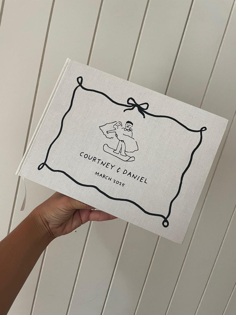 May include: A linen-covered wedding photo album with a black line drawing of a couple on a snowboard. The album is inscribed with "COURTNEY & DANIEL MARCH 2025" in black text. A pink ribbon bookmark is visible.