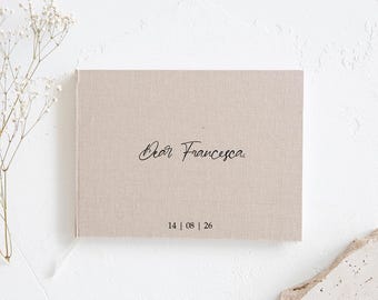 May include: A beige linen-covered book with the words "Dear Francesca" in elegant script. The date "14 | 08 | 26" is printed below the text. The book is on a white surface with dried flowers.