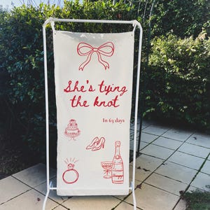 May include: A white sign with red illustrations and text. The sign reads "She's tying the knot" with drawings of a bow, cake, shoes, champagne, and a ring. The text "In 63 days" is also present.