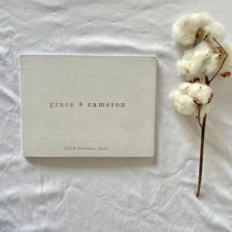 Op de afbeelding: Een lichtgekleurd, rechthoekig boek met de namen "grace + cameron" en de datum "22nd October 2022" op de kaft. Een takje katoen met pluizige witte bloemen is rechts van het boek geplaatst. Het boek ligt op een witte stoffen ondergrond.