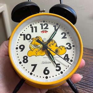 May include: A vintage yellow Garfield alarm clock with black bells and legs. The clock face features the Garfield character and the Sunbeam logo. The clock hands are black with a green second hand.