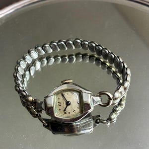 May include: Vintage silver-toned wristwatch with a rectangular face and a linked band. The watch face has black numerals and hands against a cream-colored background. The watch is displayed on a reflective surface.