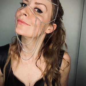 May include: A woman wearing a silver chain headpiece that covers her forehead, eyes, and nose. The chain is layered and has multiple strands. The woman is wearing a black top and has long, wavy brown hair.