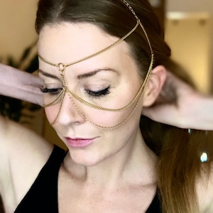 May include: A gold chain headpiece with multiple strands, worn as a headband and draped over the face. The chain is made of small, linked rings.
