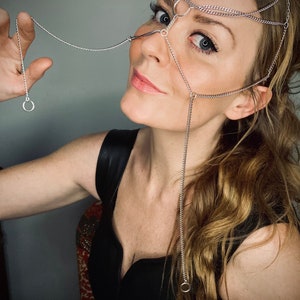 May include: Silver-toned face chain with multiple strands and small rings. The jewelry piece is designed to be worn across the forehead, over the nose, and around the face. The model is wearing a black top.