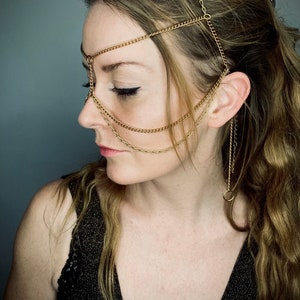 May include: A woman wearing a gold chain headpiece that covers her forehead, nose, and ears. The chain is made of multiple strands and has a delicate, feminine design. The woman is wearing a black top and has long, flowing hair.