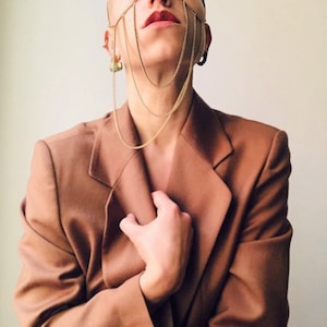 May include: A person wearing a brown blazer with a gold chain face mask. The chain mask is made of multiple strands of chain that are connected to form a geometric pattern.