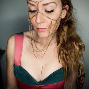 May include: A woman wearing a gold chain headpiece and a green and pink dress. The chain headpiece is a delicate design with multiple strands of chain.